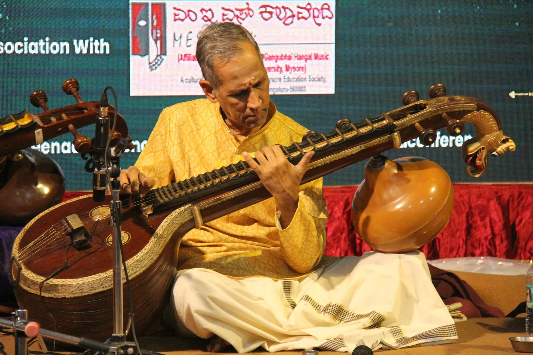 A senior vainika in a yellow kurta playing the Saraswati veena on the VTP VI stage at MES Kalavedi