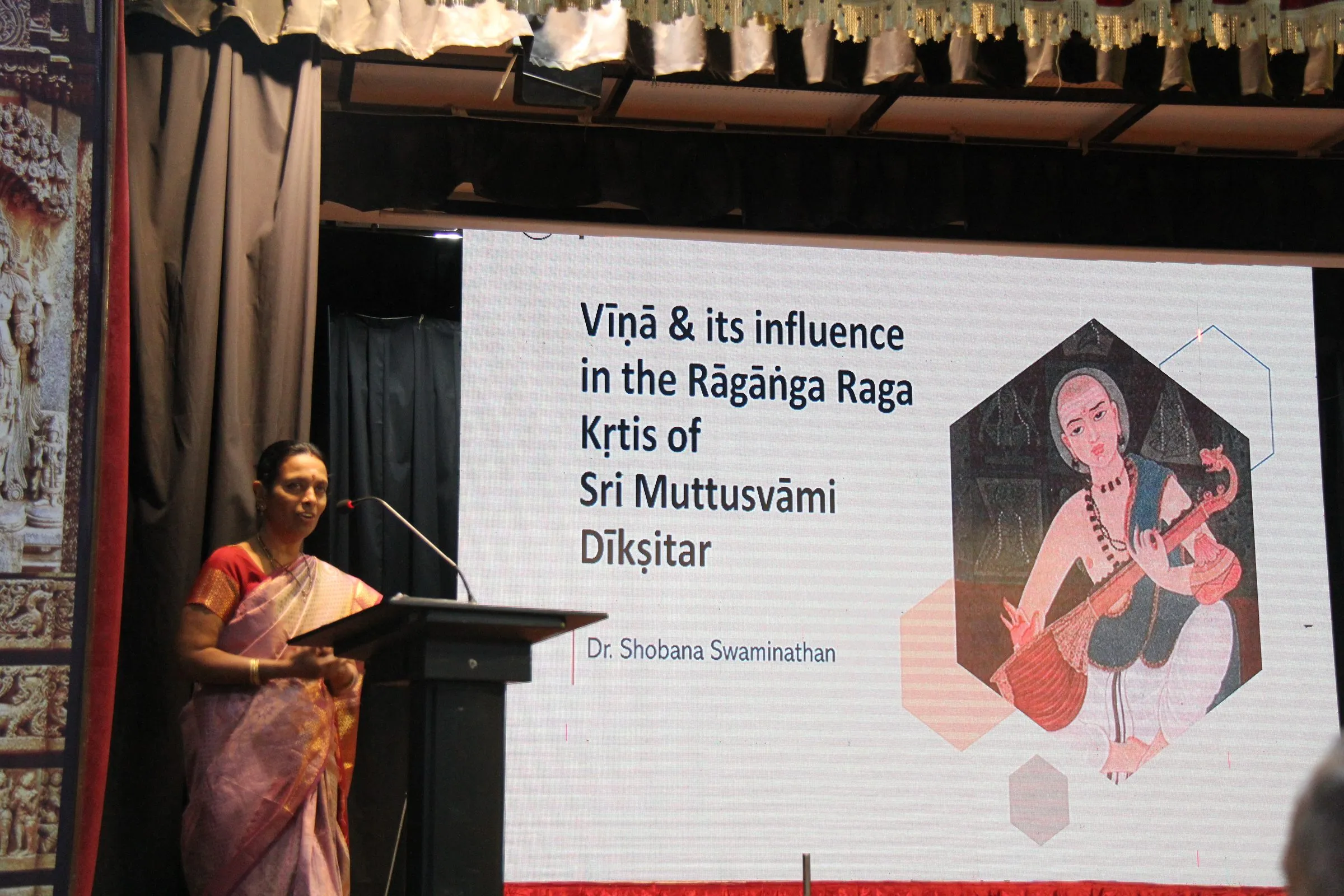 A speaker at the lectern beside a slide titled “Vīṇā & its influence in the Rāgāṅga Raga Kṛtis of Sri Muttusvāmi Dīkṣitar”